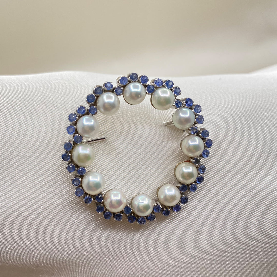 Vintage Brosche aus 750er Weißgold mit zwölf Akoya-Zuchtperlen und facettierten blauen Saphiren in klassischer Anordnung.